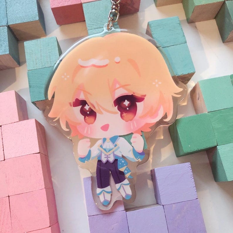 May include: A clear acrylic keychain depicting a chibi anime character with peach-coloured hair and large red eyes. The character is dressed in a white and blue outfit with purple trousers. The keychain has a silver metal ring.