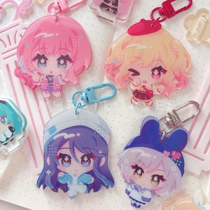May include: A collection of acrylic keychains featuring chibi-style anime characters with vibrant hair colors including pink, blonde, purple, and blue. Each charm includes a metal or plastic clasp, with some designs wearing hats or accessories.