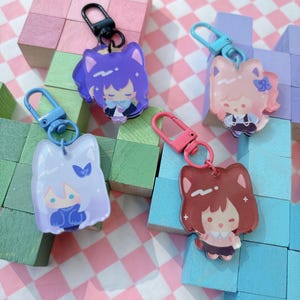 May include: Four acrylic keychains with cartoon character designs. Each keychain features a different character with a cat-like appearance, in shades of purple, pink, white, and brown. The keychains have colored clasps and are set against a backdrop of pastel-colored blocks.