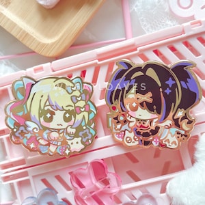 May include: Two enamel pins with cartoon character designs. One pin features a blonde character with a pink bow, the other a dark-haired character with sunglasses. Both pins have gold-colored outlines and colorful details.