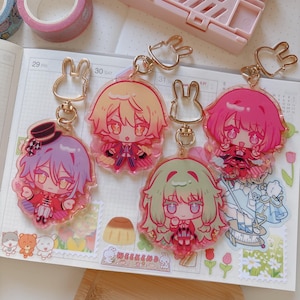 May include: Five anime-style character keychains with gold-colored bunny-shaped clasps. The characters have different hair colors: purple, yellow, pink, and green. The keychains are on a planner with stickers and pink washi tape.