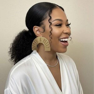 May include: Gold-colored, arched earrings with the text "PRETTY & PETTY" engraved on them. The earrings are worn by a person wearing a white robe and a silver necklace. The person is smiling.