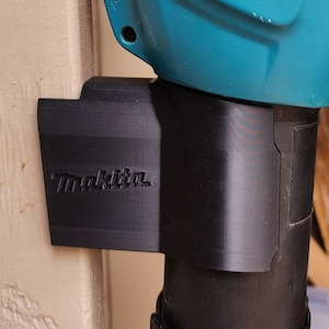 May include: A black plastic wall mount for a leaf blower with the word "Makita" printed on it.