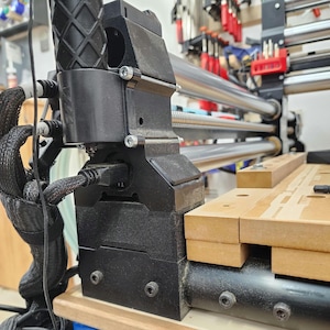 Peut inclure: Gros plan sur une imprimante 3D noire avec une poignée noire et une tige métallique argentée. L'imprimante est montée sur un établi en bois.