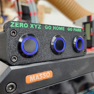 May include: A black control panel with three blue illuminated push buttons labeled "ZERO XYZ GO HOME GO PARK". The panel is mounted on a black surface with the word "MASSO" printed in orange.