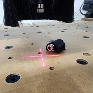 May include: A black laser module projects a bright pink crosshair onto a light brown wooden surface. The laser is mounted on a CNC machine, with a black brush head visible above. The surface has several circular holes.