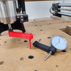 May include: A red plastic depth gauge with the text "Dash Made Woodworking" and a black dial indicator gauge with a silver face. The gauges are on a wooden surface with a pattern of holes.