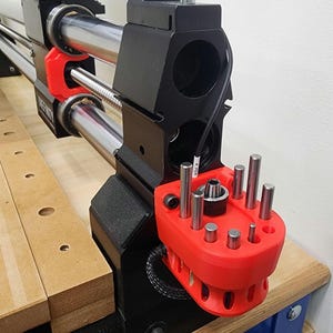 May include: Close-up of a black and red CNC machine component. The machine features silver rods, a red tool holder with multiple silver pins, and a black cable. The component is mounted on a wooden surface.