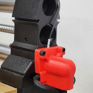 Może przedstawiać: Czerwona obudowa 3D do drukarki 3D, z czarnym kablem i czarnym plecionym kablem wychodzącym z niej. Obudowa jest przymocowana do czarnej metalowej ramy za pomocą dwóch srebrnych śrub.