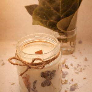 Puede incluir: Una vela en tarro de cristal transparente con una mecha de madera y un lazo de cuerda. La vela está rellena de cera cremosa y decorada con flores y hojas secas. Un pequeño tarro de cristal con hojas verdes está en el fondo.