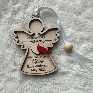 May include: Wooden angel ornament with the text "CARRIED FOR A moment AND LOVED FOR A lifetime Baby Anderson May 2023". Features a red cardinal and a white ribbon for hanging.