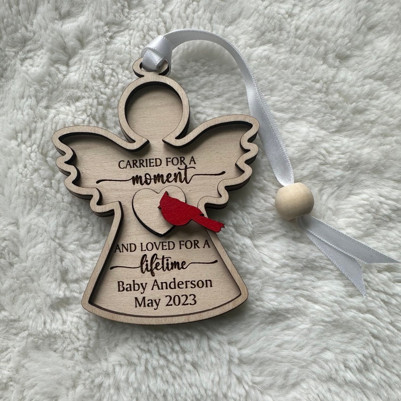 May include: Wooden angel ornament with the text "CARRIED FOR A moment AND LOVED FOR A lifetime Baby Anderson May 2023". Features a red cardinal and a white ribbon for hanging.