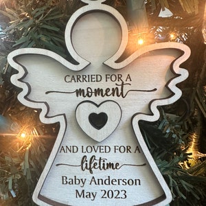 May include: Wooden angel ornament with the text "CARRIED FOR A moment AND LOVED FOR A lifetime Baby Anderson May 2023." The ornament has a heart cutout and a teal ribbon for hanging.