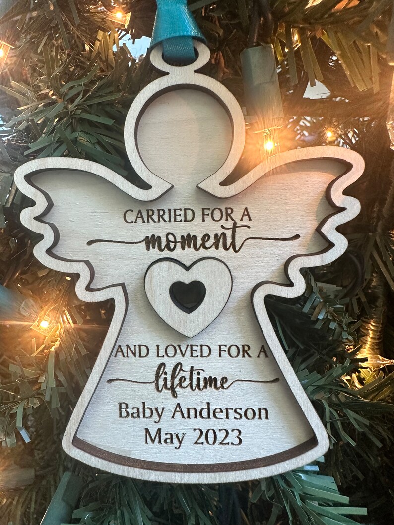May include: Wooden angel ornament with the text "CARRIED FOR A moment AND LOVED FOR A lifetime Baby Anderson May 2023." The ornament has a heart cutout and a teal ribbon for hanging.