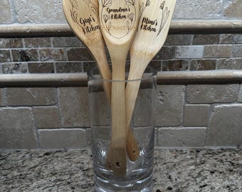 May include: Three wooden spoons with floral designs and personalized text. The spoons are in a glass jar on a granite countertop. The text on the spoons reads "Gigi's Kitchen", "Grandma's Kitchen", and "Mimi's Kitchen".