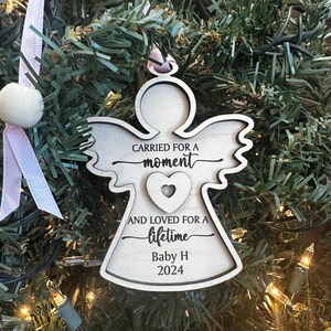 May include: Wooden angel Christmas ornament with the text "CARRIED FOR A moment AND LOVED FOR A lifetime Baby H 2024". The ornament is hanging on a Christmas tree with pink ribbon and a white pom-pom.
