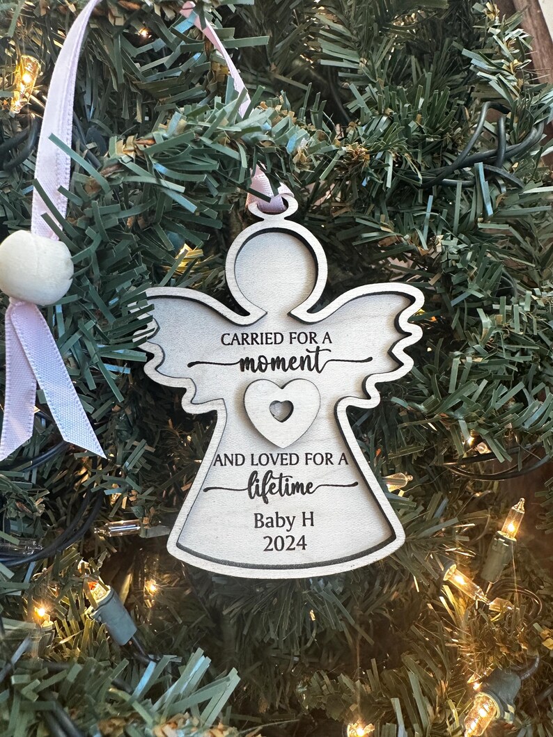 May include: Wooden angel Christmas ornament with the text "CARRIED FOR A moment AND LOVED FOR A lifetime Baby H 2024". The ornament is hanging on a Christmas tree with pink ribbon and a white pom-pom.