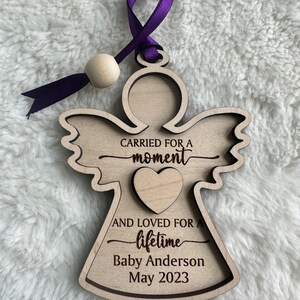 May include: Wooden angel ornament with a purple ribbon and a wooden bead. The angel has the text "Carried for a moment and loved for a lifetime" with the name "Baby Anderson" and the date "May 2023".