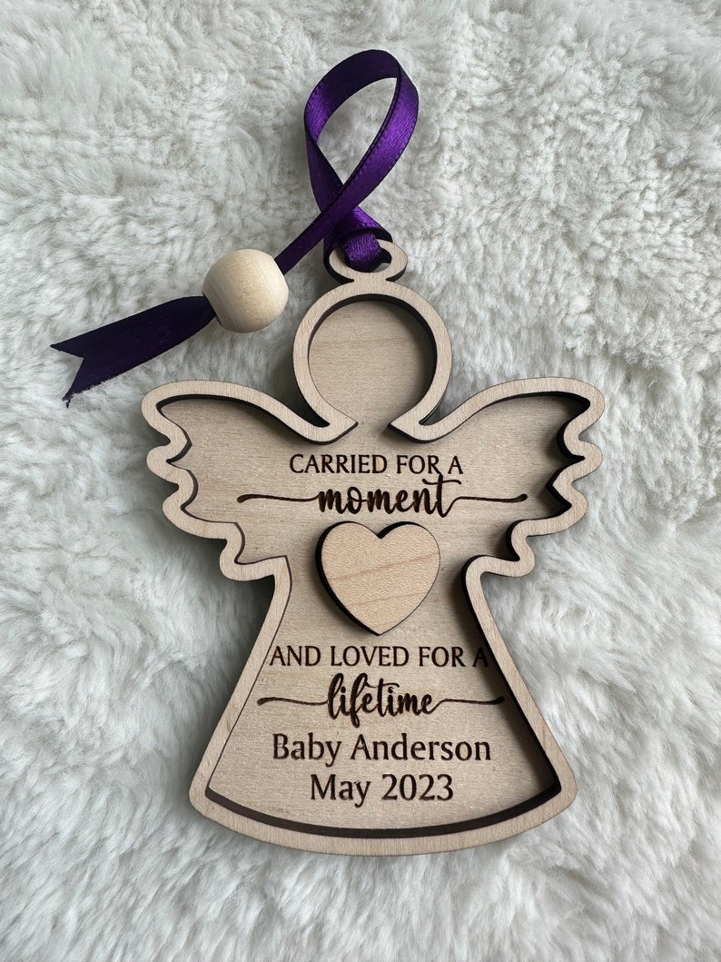 May include: Wooden angel ornament with a purple ribbon and a wooden bead. The angel has the text "Carried for a moment and loved for a lifetime" with the name "Baby Anderson" and the date "May 2023".