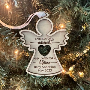 May include: A wooden angel ornament with a heart-shaped cutout and the text "Carried for a moment and loved for a lifetime." Includes the name "Baby Anderson" and the date "May 2023". The ornament hangs from a white ribbon.