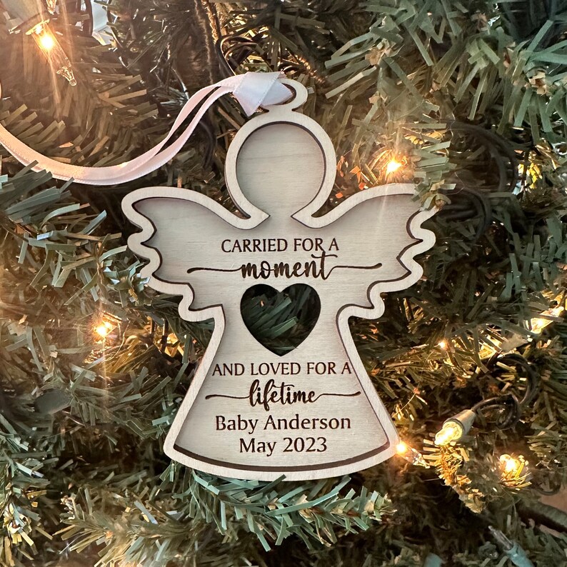 May include: A wooden angel ornament with a heart-shaped cutout and the text "Carried for a moment and loved for a lifetime." Includes the name "Baby Anderson" and the date "May 2023". The ornament hangs from a white ribbon.