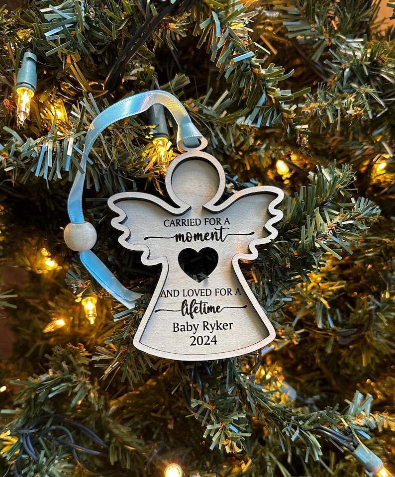 May include: Wooden angel Christmas ornament with the text "CARRIED FOR A moment AND LOVED FOR A lifetime Baby Ryker 2024". The ornament has a heart cutout and is suspended by a light blue ribbon.