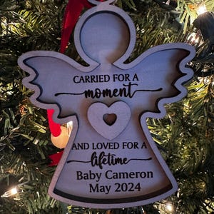 May include: Wooden angel Christmas ornament with the text "CARRIED FOR A moment AND LOVED FOR A lifetime Baby Cameron May 2024". The angel has a heart cutout and is hanging from a red ribbon.
