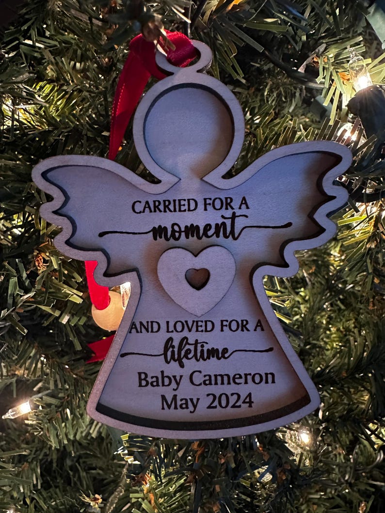 May include: Wooden angel Christmas ornament with the text "CARRIED FOR A moment AND LOVED FOR A lifetime Baby Cameron May 2024". The angel has a heart cutout and is hanging from a red ribbon.