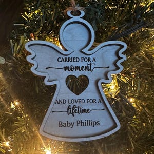 May include: A wooden angel ornament with the text "CARRIED FOR A moment AND LOVED FOR A lifetime Baby Phillips." The angel has a heart-shaped cutout and is hanging from a string. The ornament is displayed on a Christmas tree.