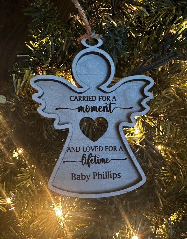 May include: A wooden angel ornament with the text "CARRIED FOR A moment AND LOVED FOR A lifetime Baby Phillips." The angel has a heart-shaped cutout and is hanging from a string. The ornament is displayed on a Christmas tree.
