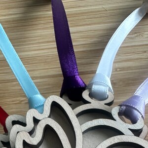 May include: Close-up of a wooden ornament with a layered design, featuring colorful satin ribbons in teal, red, purple, white, lavender, and pink. Each ribbon is attached to a wooden bead and the ornament.