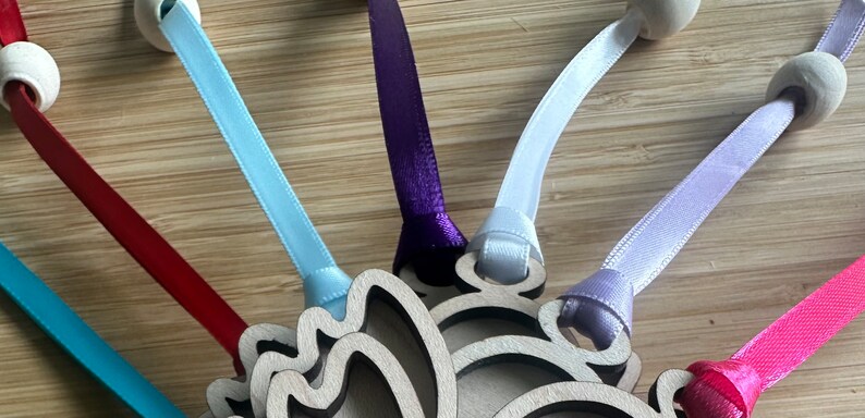 May include: Close-up of a wooden ornament with a layered design, featuring colorful satin ribbons in teal, red, purple, white, lavender, and pink. Each ribbon is attached to a wooden bead and the ornament.