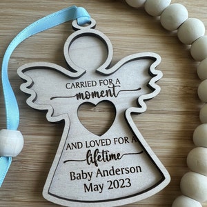 May include: A wooden angel ornament with a light blue ribbon for hanging. The angel has a heart-shaped cutout and the text "CARRIED FOR A moment AND LOVED FOR A lifetime Baby Anderson May 2023". A string of white wooden beads is in the background.