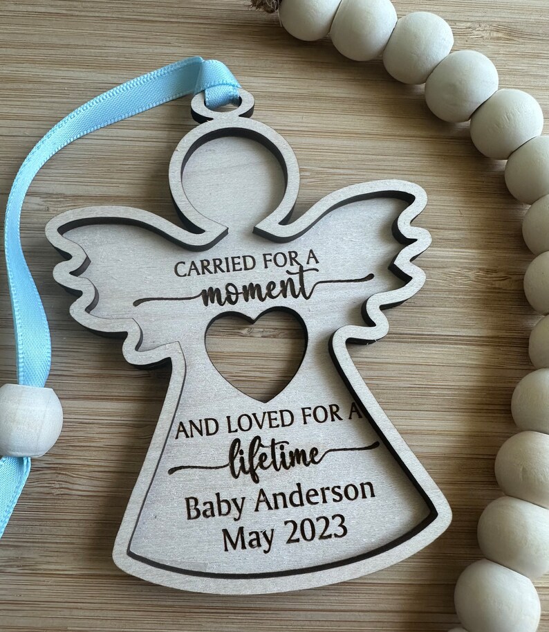 May include: A wooden angel ornament with a light blue ribbon for hanging. The angel has a heart-shaped cutout and the text "CARRIED FOR A moment AND LOVED FOR A lifetime Baby Anderson May 2023". A string of white wooden beads is in the background.