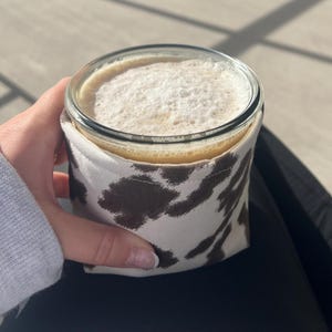 May include: A glass of creamy drink with a foamy top, held in a white and brown cow print cosy. The cosy is wrapped around the glass. The drink is in a clear glass, providing insulation and a decorative touch.