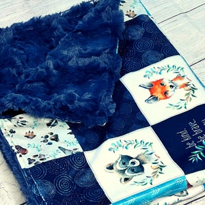May include: A blue and white baby blanket with a patchwork design featuring woodland animals, including a fox, raccoon, and paw prints. The blanket has a soft, fuzzy texture and includes the text "Be kind, be brave, be you".