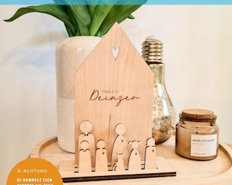 May include: A wooden house-shaped sign with the text "Familie Deinzer" and cutouts for a family of five, including a cat. The sign is sitting on a wooden surface.
