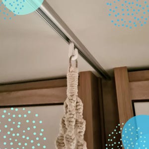 Könnte beinhalten: Ein weißer Kunststoffhaken, der an einer silbernen Schiene befestigt ist und einen beigefarbenen Makramee-Pflanzenaufhänger trägt. Das Makramee besteht aus gewebtem Seil und hängt herunter, bereit für eine Topfpflanze. Der Hintergrund umfasst Holzpaneele und eine weiße Decke.