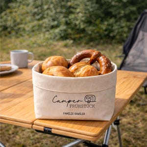 Könnte beinhalten: Ein beiger Canvas-Korb gefüllt mit Brötchen und einer Brezel steht auf einem Holztisch. Auf dem Korb stehen die Worte "Camper Frühstück" und "Familie Kausler". Eine Tasse und ein Teller sind im Hintergrund.