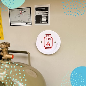 Könnte beinhalten: Ein weißer, kreisförmiger Gasbehälter mit dem Wort "GAS" und einem Flammensymbol in Rot. Der Halter ist an einer hellen Wand montiert, mit einem Gastank in der unteren linken Ecke. Es gibt auch zwei Informationstafeln.