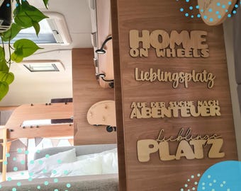 Op de afbeelding: Houten wandkunst met de zinnen "HOME ON WHEELS", "Lieblingsplatz", "AUF DER SUCHE NACH ABENTEUER" en "Lieblings PLATZ" in een camperinterieur. De kunst is gemaakt van lichtgekleurd hout en gemonteerd op een houten muur.