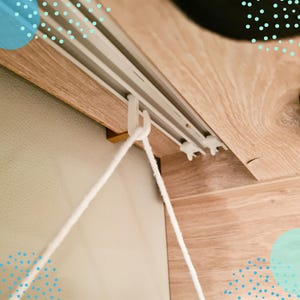 Könnte beinhalten: Nahaufnahme eines weißen Gardinenstangensystems mit einer weißen Schnur, die herunterhängt. Die Stange ist auf einer hellen Holzoberfläche montiert. Die Wand ist hellbeige.