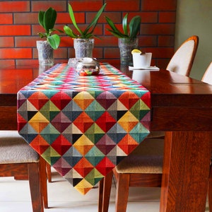 以下が含まれることがあります： 赤、青、緑、黄、茶色の三角形のパターンが施されたカラフルな幾何学模様のテーブルランナー。ランナーは木製のテーブルに置かれ、2つの椅子が置かれています。