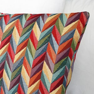 Może przedstawiać: Kolorowa poduszka dekoracyjna z geometrycznym wzorem chevron w odcieniach czerwieni, pomarańczy, żółci, zieleni, błękitu i fioletu.