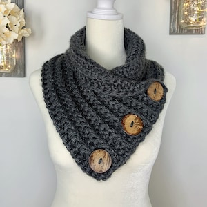 May include: A gray knitted infinity scarf with three wooden buttons. The scarf is displayed on a white mannequin.