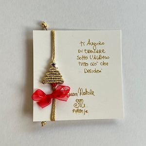 Può includere: Un biglietto di Natale con testo dorato e un piccolo ornamento a forma di campana. Il biglietto ha un fiocco rosso e un cordino dorato. Il testo sul biglietto recita "Ti Auguro di trovare sotto l'albero tutto ciò che desideri. Buon Natale Firenze."