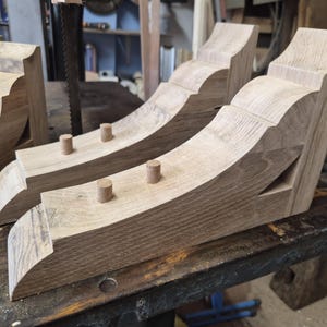 Puede incluir: Dos ménsulas de madera sin terminar en color marrón claro con un perfil decorativo curvo y una textura de grano suave. Cada ménsula tiene dos pequeñas espigas cilíndricas de madera que sobresalen de la superficie superior.