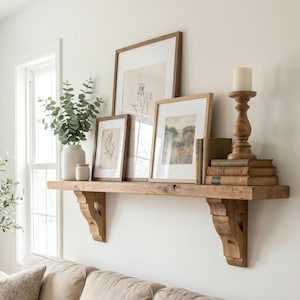 May include: A wooden wall shelf with two carved corbel brackets holding a white ceramic vase with green eucalyptus stems, three framed botanical prints, a stack of three vintage books, and a turned wooden candle holder with a white pillar candle.