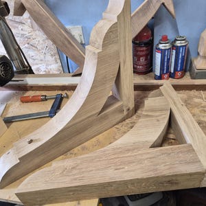 May include: Wooden table legs in various stages of construction. The unfinished oak legs are light brown and feature curved and angled designs. Tools and materials, including a clamp and spray cans, are visible in the background, suggesting a woodworking workshop.