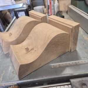 May include: Close-up of unfinished wooden furniture components. Two curved, wave-shaped pieces and two rectangular blocks are visible. The wood has a natural grain and is light brown in color. A metal ruler is in the foreground.